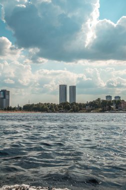 Tarihi bir şehir silueti ve arka planında tüylü beyaz bulutlarla nehirde devriye gezen iki askeri hız teknesi.