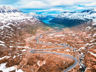 Karla kaplı dağlar, kayalık arazi ve akan bir nehir ile çevrili İzlanda 'daki geniş bir vadiden geçen dolambaçlı bir yolun nefes kesici hava manzarası..