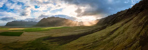 Stock image Breathtaking aerial view of Iceland's majestic landscape with lush green fields, dramatic mountains, and sunrays piercing through clouds, showcasing the beauty of this travel destination.