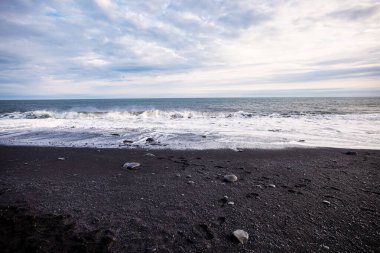 Dalgalar, İzlanda 'nın Reynisfjara sahillerinde kara kumlara çarpıyor. Gökyüzü bulutlu.