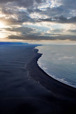 İzlanda 'da gün batımında, dramatik bulutlu bir gökyüzü altında Atlantik Okyanusu' yla buluşan siyah kumsal manzarası.