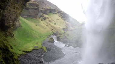 İzlanda 'daki Kvernufoss şelalesinin arkasından bakıldığında, yemyeşil yosun, engebeli kaya oluşumları ve resim gibi bir manzarada çağlayan bir şelale ile gizli bir mücevher sergilenmektedir.