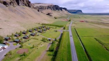 İzlanda kırsalında yuva yapmış, göz kamaştırıcı uçurumların arka planına kurulmuş resimli kulübelerin havadan görünüşü.