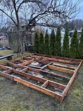 Arka planda yeşil ağaçlar ve bulutlu gökyüzü manzaralı bir arka bahçe yapısı için ahşap bir temel inşa ediliyor.