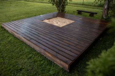 Ağaçlar ve çalılarla çevrili arka bahçesinde merkezi bir kum havuzu olan yeni boyanmış ahşap güverte.