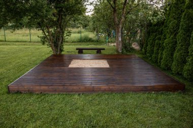 Ağaçlar ve doğal çevreyle çevrili, arka bahçesinde merkezi kum havuzu olan ahşap güverte.