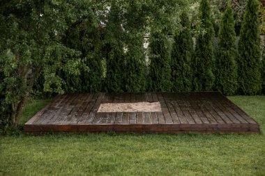 Ağaçlar ve çalılarla çevrili arka bahçesinde merkezi bir kum havuzu olan yeni boyanmış ahşap güverte.