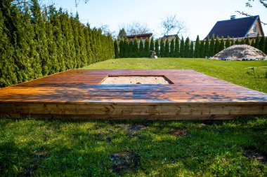 Ağaçlar ve çalılarla çevrili arka bahçesinde merkezi bir kum havuzu olan yeni boyanmış ahşap güverte.