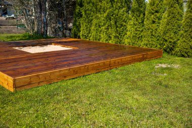 Ağaçlar ve çalılarla çevrili arka bahçesinde merkezi bir kum havuzu olan yeni boyanmış ahşap güverte.