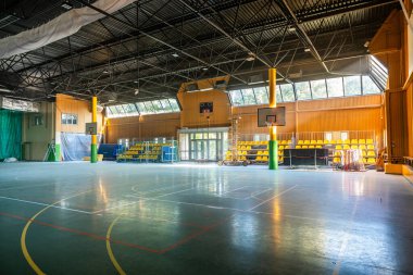 Geniş bir spor salonu. Basketbol sahası, oturma ve ekipmanı var. Büyük pencerelerden güneş ışığıyla yıkanmış..