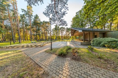 Ağaçlık bir parktaki açık hava amfi tiyatrosu. Sahneye bakan sıra sıra dizilmiş uzun banklar. Hepsi de uzun ağaçların altında..