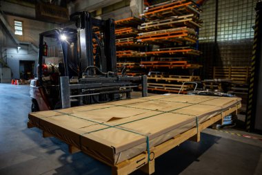 Bir forklift büyük bir deponun içinde çalışır, büyük, ahşap paletlere sarılı bir paket taşır, depolama raflarıyla çevrilidir..