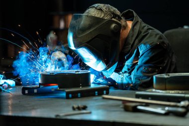 Welder creating a bright arc while joining metal pieces in a dark workshop, surrounded by tools and equipment. clipart