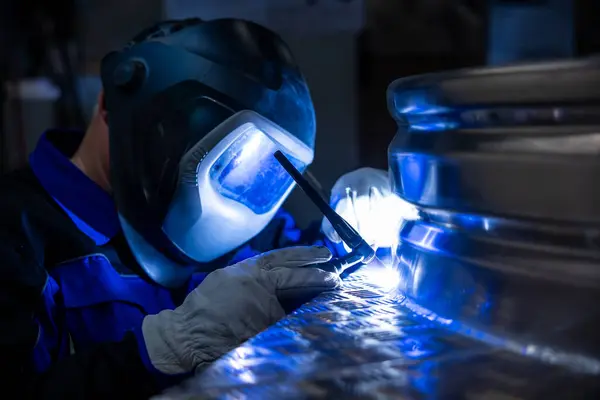 Koruyucu teçhizattaki kaynakçı, loş aydınlatılmış bir atölyede metal bir yapıyı kaynak yapmaya odaklanmış, kaynak sürecini vurguluyor.