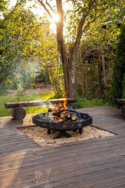 Ahşap bir güvertede, yemyeşil ve yüksek yeşilliklerle çevrili yanan odunları olan dairesel bir ateş çukuru bulunur. Kırsal banklar dingin ayarları artırır.