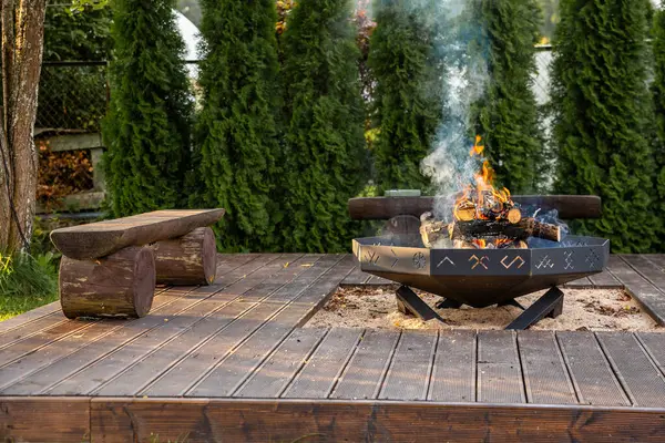 Ahşap bir güvertede yanan odunlar ve dumanlı dairesel bir ateş çukuru bulunur. Yeşillik ve yüksek yeşilliklerle çevrili kırsal banklar manzarayı güzelleştiriyor..