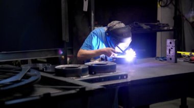 Koruyucu dişli bir işçi, etkin bir endüstriyel ortamda ekipman ve parçalarla çevrili bir tezgahta metal kaynaklıyor..