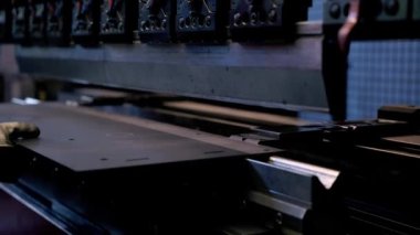 Metal levhaları şekillendirmek için klempleri ve kolları olan büyük bir endüstriyel makine kullanılır. Bir işçinin eldivenli eli loş ışıkta elle çalıştırılmasını önerir.