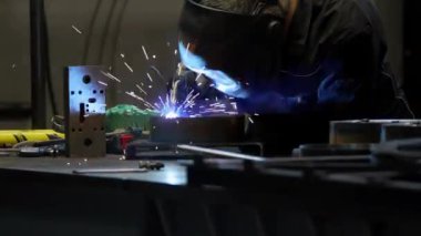 Koruyucu dişli bir işçi, malzeme ve parçalarla dolu endüstriyel atölyeyi aydınlatan parlak kıvılcımlarla bir tezgahta metal kaynaklıyor..