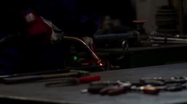 Bir kaynakçı meşale kullanır ve metal üretimi endüstrisinde parlak mavi kıvılcımlar yaratır. Endüstriyel ekipman ve araçlar arka planda görünür.