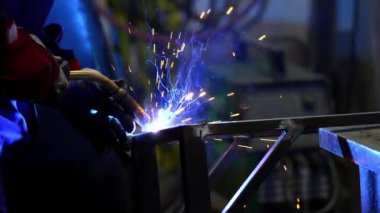 Mavi iş kıyafetli bir kaynakçı kaynak makinesi kullanır ve metal imalatında parlak kıvılcımlar yaratır. Sahne kaynak yayının mavi ışığıyla aydınlanıyor..