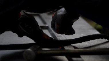 Kaynakçının eldivenli elleri bir meşale tutuyor, mavi ışık ve duman yayıyor. Metal yüzey ve çekiç görünür olup, metal işleme sürecini vurgular..