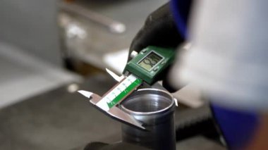 Siyah eldivenli bir kişi metal silindirin çapını ölçmek için dijital kalibre kullanır, bir imalat ayarında hassasiyet ve doğruluğu vurgular..