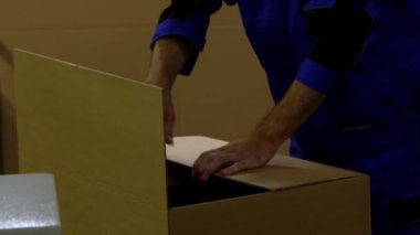 Mavi üniformalı biri, karton bir kutuyu mühürlemek için el bilgisayarı kullanıyor. Sanayi ortamında diğer kutular ve beton bir duvar yer alıyor.