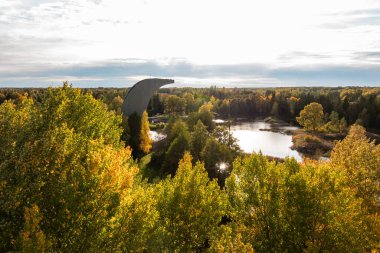 Altın ve yeşil yapraklı yemyeşil bir ormanda eşsiz bir kavisli gözlem kulesi bulunuyor. Sakin bir su kütlesi, sıcak bir günbatımı parıltısının altında gökyüzünü yansıtır..