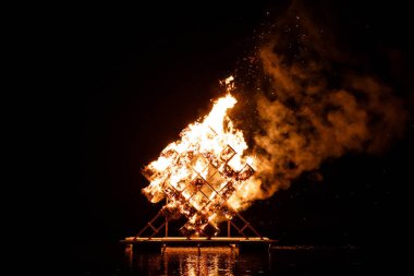 Geometrik ahşap bir yapı, suyun üzerindeki platformda parlak bir şekilde yanar. Yansımalar yapar ve gece gökyüzünü yoğun alevlerle aydınlatır..