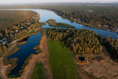 Kıvrımlı bir nehir sık ormanlar ve açık araziler boyunca akar, kıyılar boyunca küçük ev kümeleri kırsal bir alanı işaret eder..