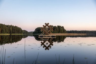 Yoğun ormanlarla çevrili sakin bir göl, geometrik ahşap bir yapıya sahiptir. Açık gökyüzü maviden pembeye, suyun üzerinde nilüfer yaprağı var..