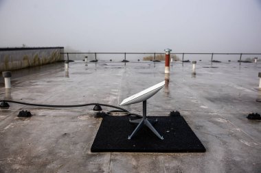 A flat rooftop features a satellite dish on a black mat, surrounded by fog. The modern building's railing is visible, with muted colors and a damp surface. clipart