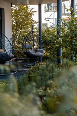 Açık teras alanında yoğun yeşilliklerle çevrili iki tane asılmış yumurta koltuğu bulunur. Arka planda balkonlu bir bina görülebilir.