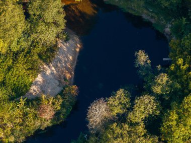 Hava perspektifi, koyu suyla çevrili küçük, dolambaçlı bir nehri, canlı yeşil yapraklarla çevrili ve sol tarafında kumlu bir sahili yakalar..