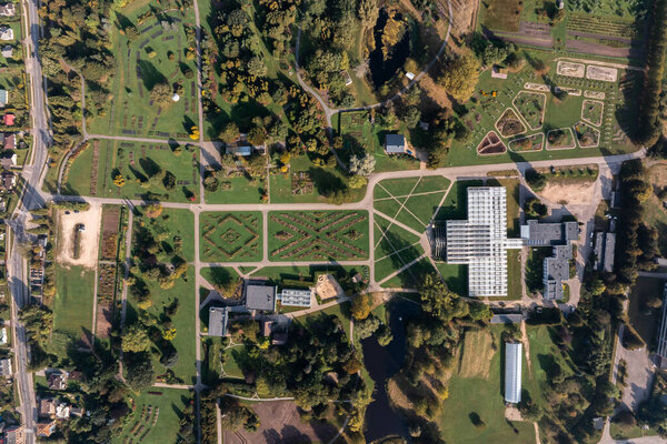 An aerial view captures a botanical garden's geometric landscape, featuring a large greenhouse, tree lined paths, and diverse garden sections in an urban setting.