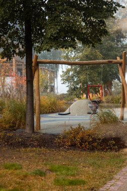 Bir oyun parkında, sonbahar yapraklı ağaçlarla çevrili ahşap bir salıncak ve kum tepeciği bulunur. Park alanından geçen bir patika var..