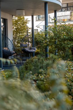 Çağdaş bir terasta, bir kentsel yerleşim alanındaki balkon ve pencerelerin bir zeminine dayanan bol yeşillikler arasında asılı iki yumurta sandalyesi yer alıyor..