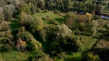 Geniş bir botanik bahçesinde yeşil ve sonbahar renklerinde çeşitli ağaçlar ve çalılıklar, kıvrımlı yollar ve arka planda görünen bir şehir manzarası bulunur..