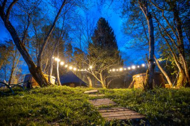 Yıldız patlaması ışıkları altında hamakları olan büyülü orman kamp alanı, yüksek ağaçlar ve gece gökyüzü arasında kurulmuş sakin bir açık hava inzivası.