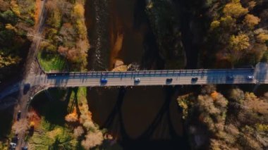 Letonya 'nın Sigulda kentindeki bir köprünün hava perspektifi, nehirden geçen araçlar. Sahne canlı sonbahar renklerinde bir orman tarafından çerçevelenmiştir..