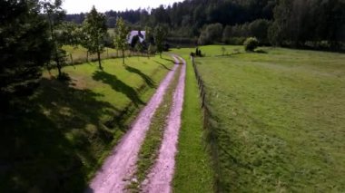 Huzurlu bir kırsaldan geçen toprak bir yol, yemyeşil çimenler ve genç ağaçlarla çevrili, ormanın yakınındaki rahat bir eve doğru ilerliyor..