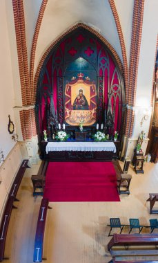 Görüntü, Gotik tarzı kemerlere karşı dizilmiş dini bir ikon, çiçek aranjmanları ve mumlarla süslenmiş zengin bir sunağın yer aldığı bir kiliseyi gösteriyor..