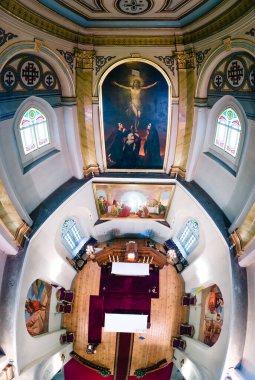Görüntü bir katedralin içini gösteriyor. Karmaşık tavan desenleri, çarmıha germe resmi, boyalı cam ve simetrik sıralar var..