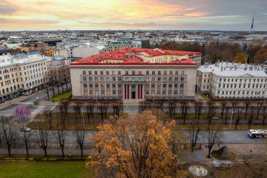 Riga, Letonya 'da kırmızı çatılı ve simetrik tasarımlı önemli bir binanın havadan görünüşü. Riga TV Kulesi sıcak bir arka planda görünür..