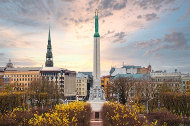 Özgürlük Anıtı Riga, Letonya 'da duruyor, tarihi binalar ve St. Peter Kilisesi ile çevrili. Sonbahar ağaçları ve renkli bir gökyüzü manzarayı çerçeveliyor.
