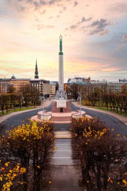 Özgürlük Anıtı, Letonya 'nın Riga şehrinde sonbahar ağaçlarıyla çevrili duruyor. Ufuk çizgisi, Aziz Peter Kilisesi kulesiyle, renkli bir günbatımına karşı..