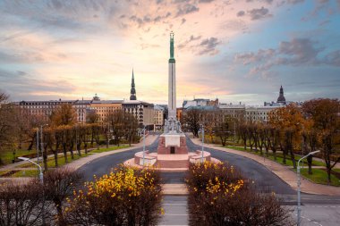 Özgürlük Anıtı, Letonya 'nın Riga şehrinde ağaçlarla çevrili bir meydanın merkezinde yer alıyor. Pastel gökyüzüne karşı tarihi yapılar ve kilise kuleleri görünür..