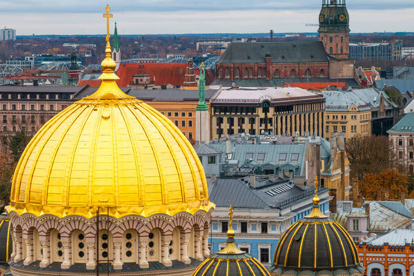 Золотой купол собора Рождества Христова выделяется на фоне Риги, под облачным небом виднеется шпиль церкви Святого Петра.