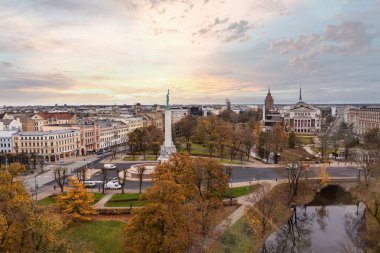 Riga, Letonya, ortada ağaçlarla çevrili Özgürlük Anıtı ve dairesel bir yol. Ufuk çizgisi tarihi ve modern mimariyi içerirken, uzakta Daugava Nehri ve sonbahar yaprakları sıcaklık katar..